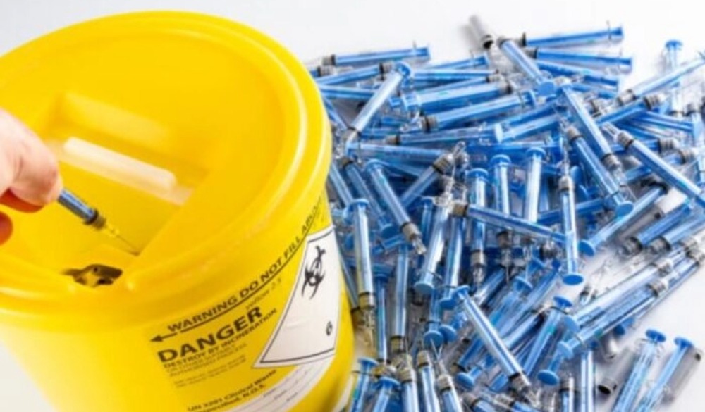 A hand safely disposes of a syringe by dropping it into a yellow sharps disposal bin. Next to the bin is a pile of blue syringes.