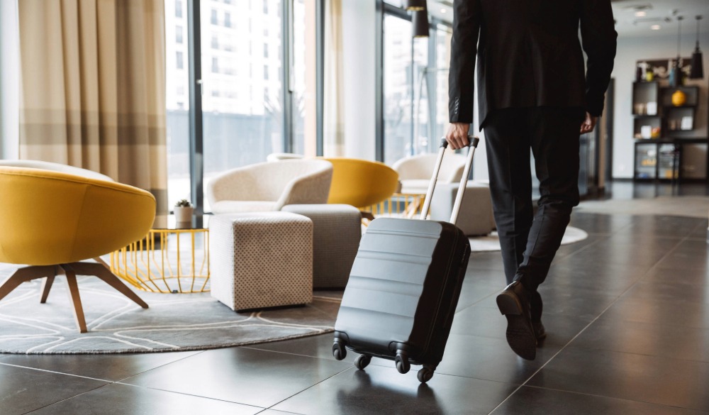 A businessman in a black suit wheels a small, grey suitcase across the floor of a contemporary hotel lobby.