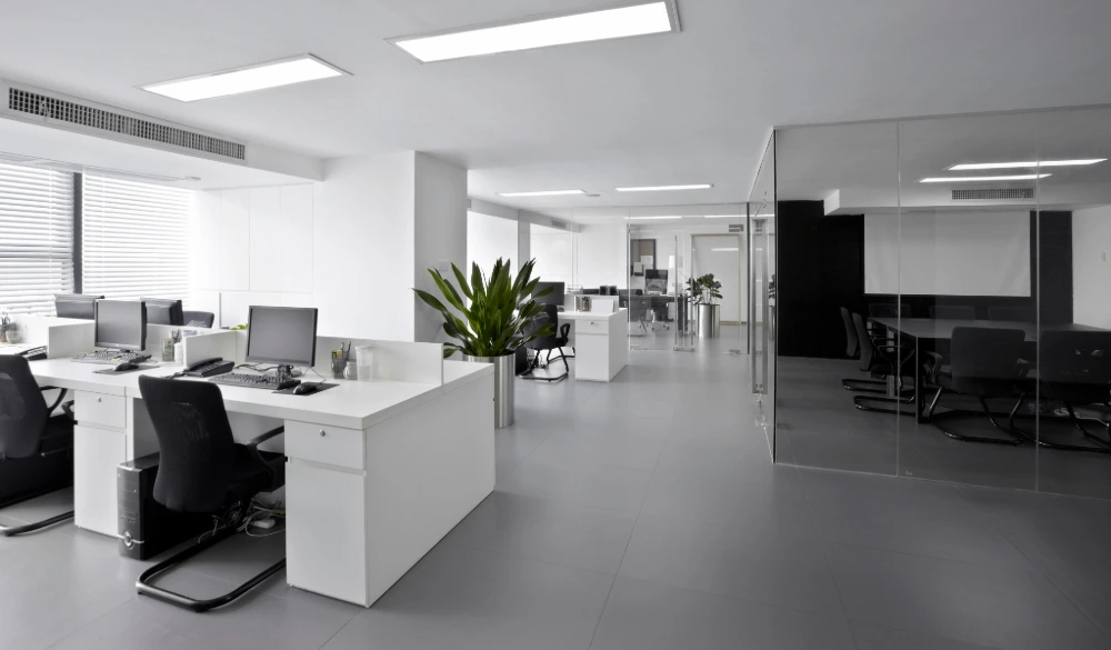 A modern office space with white desks, chairs, and walls. There are several computers on the desks, and a glass wall separates the main office area from a smaller meeting room.