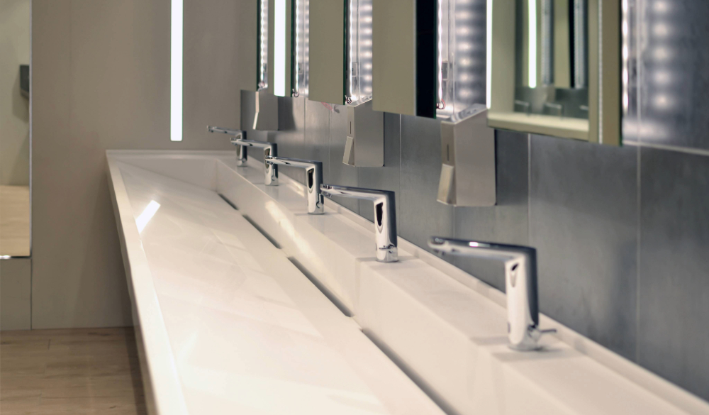 A long, white countertop in a washroom with several sinks lined up. Each sink has a silver faucet with a soap dispenser next to it. Large mirrors with bright lights hang above the sinks. 