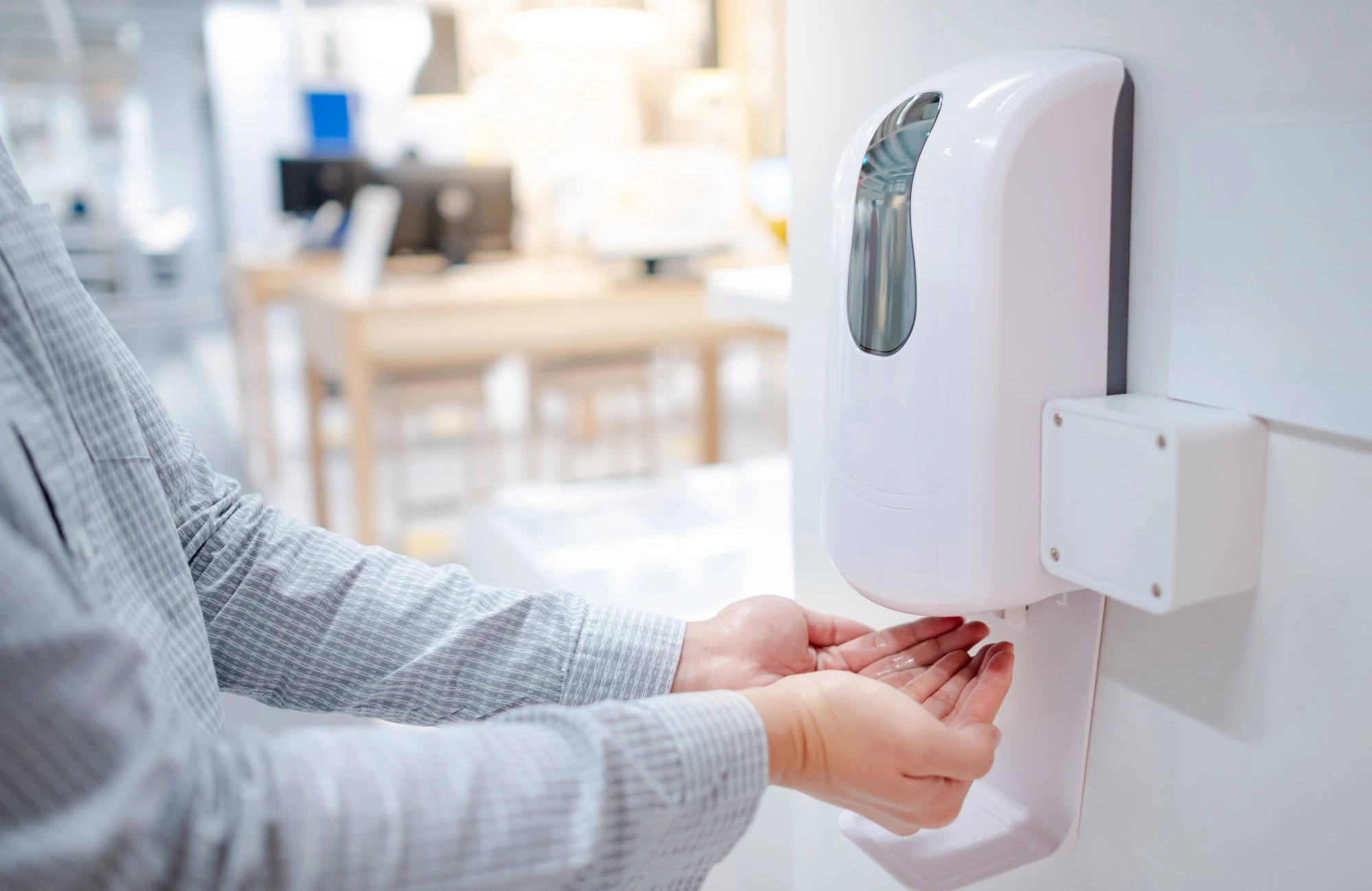 A man wearing a white and grey shirt cups his hands beneath a wall-mounted hand sanitiser dispenser in an office. 